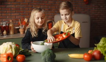 Little girl and boy mixing cutted pepper with other fresh ingredients for salad in big bowl while cooking at table with various fresh vegetables and fruits