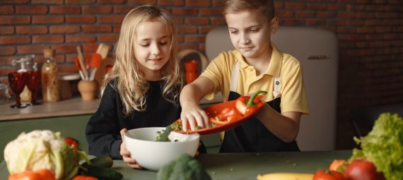 Little girl and boy mixing cutted pepper with other fresh ingredients for salad in big bowl while cooking at table with various fresh vegetables and fruits