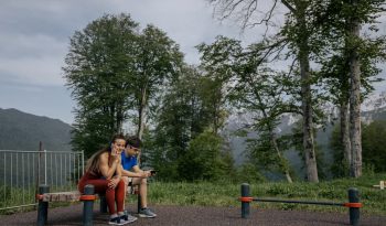 A couple in sports clothing sitting on a bench outdoors, surrounded by trees and mountains.