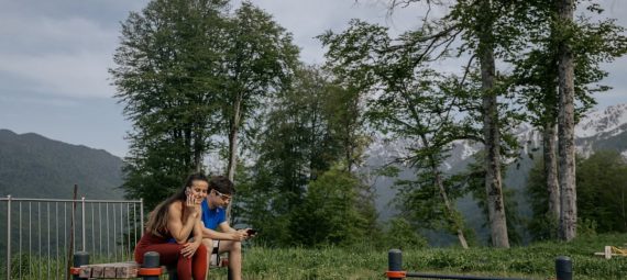 A couple in sports clothing sitting on a bench outdoors, surrounded by trees and mountains.